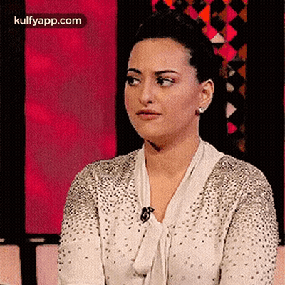 A Woman In A White Sweater With Rhinestones Is Sitting In Front Of A Pink Background . GIF