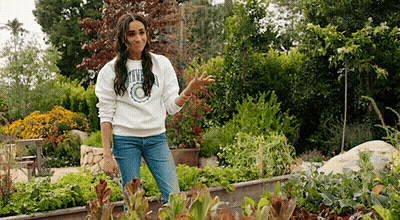 A Woman In A White Sweatshirt Is Standing In A Garden Surrounded By Plants And Flowers . GIF
