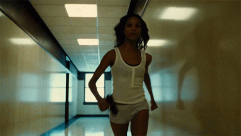 A Woman In A White Tank Top And Shorts Is Running In A Hallway GIF