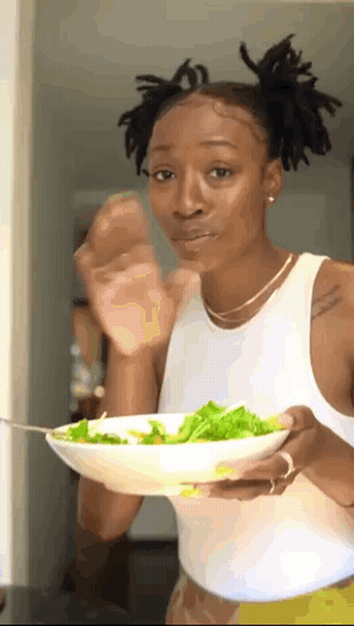 A Woman In A White Tank Top Is Holding A Bowl Of Salad With A Fork . GIF