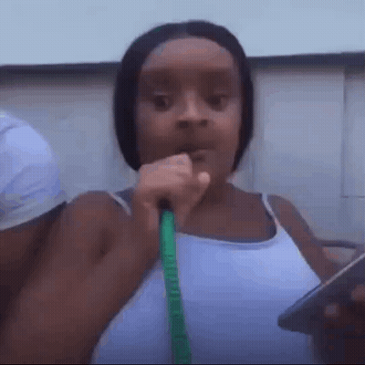 A Woman In A White Tank Top Is Holding A Green Hose In Her Mouth . GIF