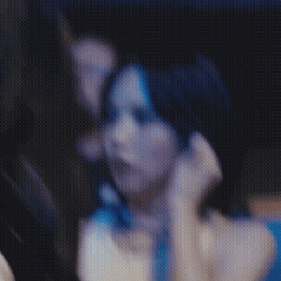 A Woman In A White Tank Top Is Sitting In A Dark Room With A Man In The Background . GIF