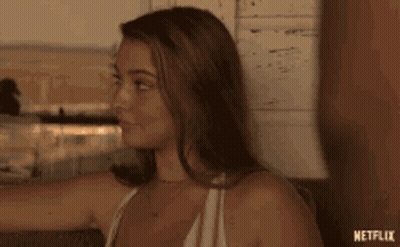 A Woman In A White Tank Top Is Sitting On A Couch And Looking At Something . GIF