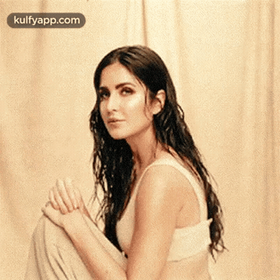 A Woman In A White Tank Top Is Sitting On The Floor With Her Legs Crossed And Looking At The Camera . GIF