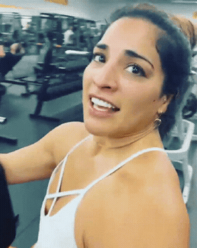 A Woman In A White Tank Top Is Smiling And Looking At The Camera GIF