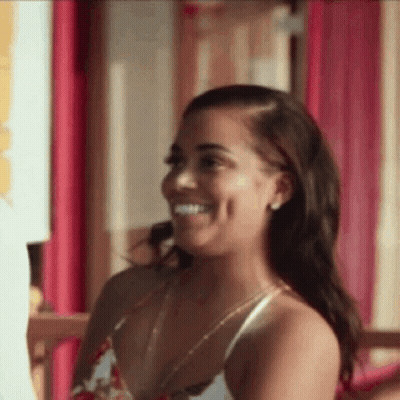 A Woman In A White Tank Top Is Smiling While Sitting In Front Of A Pink Wall . GIF