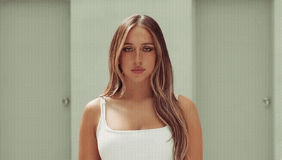 A Woman In A White Tank Top Is Standing In A Hallway Between Two Doors . GIF