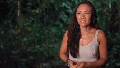 A Woman In A White Tank Top Is Standing In The Woods GIF