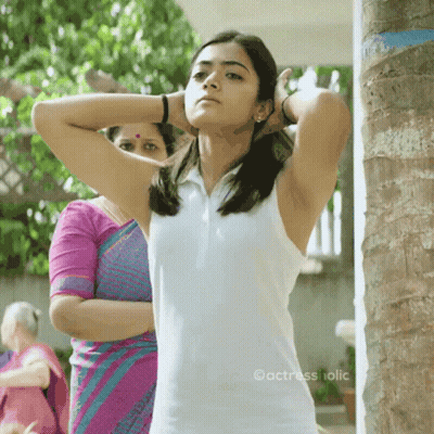 A Woman In A White Tank Top Is Standing Next To A Palm Tree . GIF