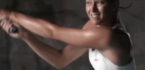 A Woman In A White Tank Top Is Swinging A Tennis Racquet . GIF