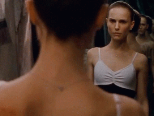 A Woman In A White Tank Top Looks At Her Reflection In A Mirror GIF