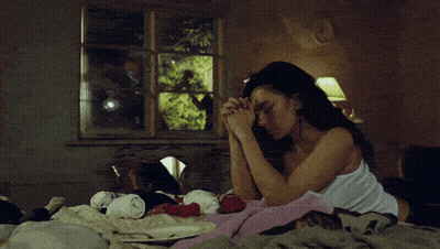 A Woman In A White Tank Top Sits On A Bed With Her Hands Folded GIF