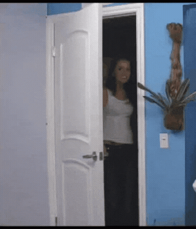A Woman In A White Tank Top Stands In A Doorway In Front Of A Blue Wall GIF