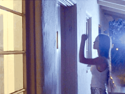 A Woman In A White Tank Top Stands In Front Of A Window GIF