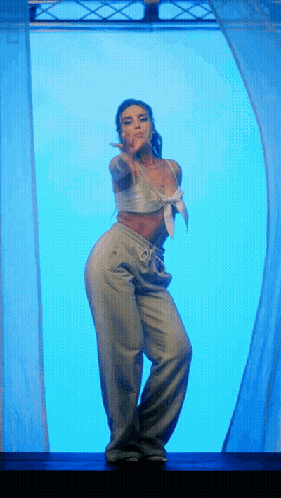 A Woman In A White Top And Grey Pants Is Dancing In Front Of A Blue Curtain GIF