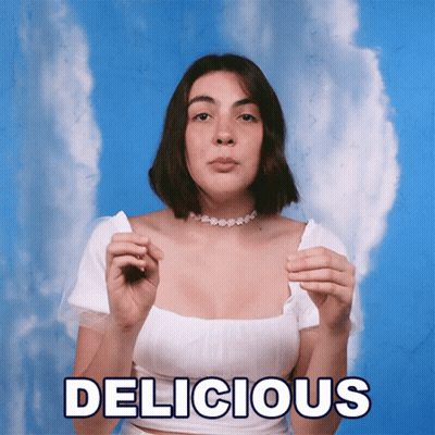 A Woman In A White Top Is Holding A Piece Of Paper With The Word Delicious Written On It GIF