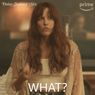 A Woman In A White Top Says " What " While Standing In Front Of A Microphone GIF