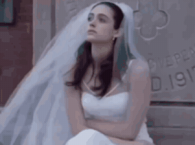 A Woman In A White Wedding Dress And Veil Is Sitting On A Bed . GIF