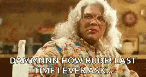 A Woman In A Wig And Glasses Is Sitting At A Table With A Spray Bottle In The Background . GIF