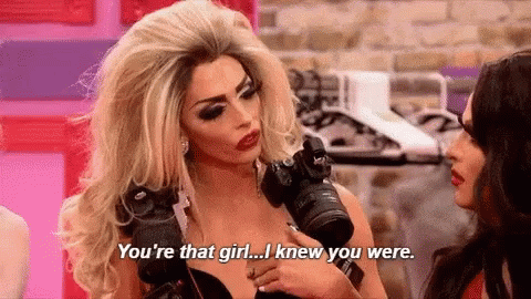 A Woman In A Wig Is Holding A Camera And Talking To Another Woman . GIF