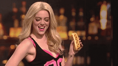 A Woman In A Wig Is Holding A Hot Dog That Says ' Vava ' On It GIF