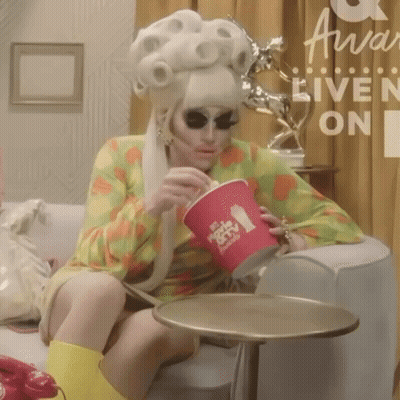 A Woman In A Wig Is Sitting On A Couch Drinking From A Cup That Says ' Movie & Tv ' On It GIF