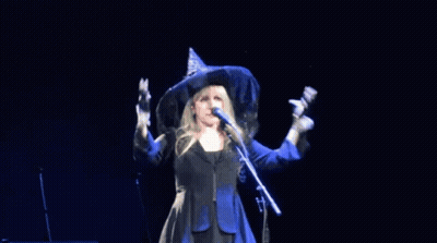 A Woman In A Witch Hat Sings Into A Microphone GIF