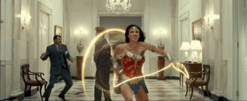 A Woman In A Wonder Woman Costume Is Holding A Lightning Bolt In A Hallway GIF