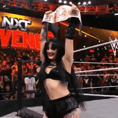 A Woman In A Wrestling Ring Holding A Championship Belt In Front Of A Sign That Says Nxt Venge GIF