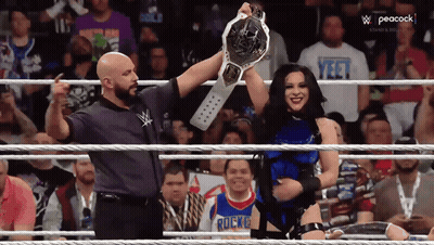 A Woman In A Wrestling Ring Holding A Championship Belt With Peacock Written On The Bottom Right GIF
