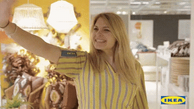 A Woman In A Yellow And Blue Striped Shirt Is Waving In Front Of A Sign That Says Ikea GIF