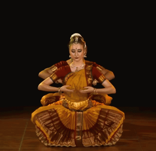 A Woman In A Yellow And Red Dress Is Sitting In A Lotus Position With Her Arms Outstretched GIF