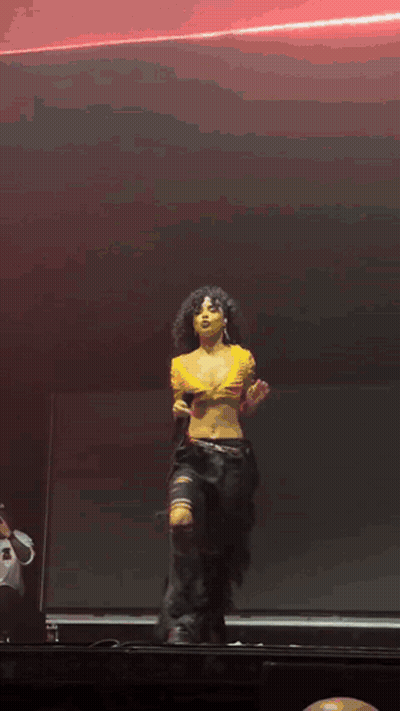 A Woman In A Yellow Crop Top And Black Pants Is Dancing On A Stage GIF