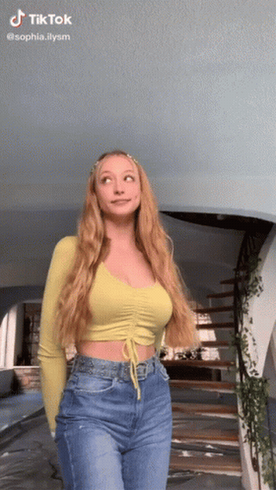 A Woman In A Yellow Crop Top And Blue Jeans Is Standing In Front Of A Spiral Staircase . GIF