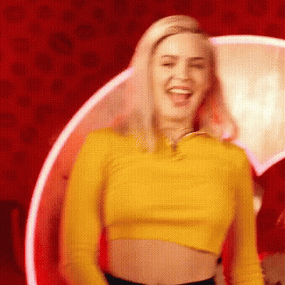 A Woman In A Yellow Crop Top Is Standing In Front Of A Red Wall And Smiling . GIF