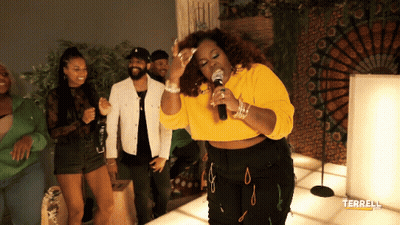 A Woman In A Yellow Crop Top Singing Into A Microphone In Front Of People GIF