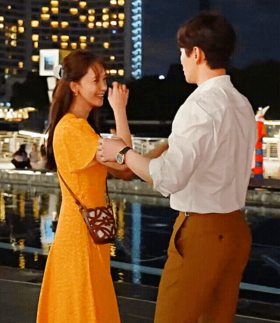 A Woman In A Yellow Dress Holds A Man 'S Hand In Front Of A Body Of Water GIF