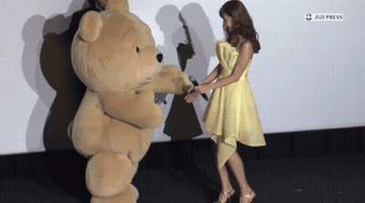 A Woman In A Yellow Dress Holds A Microphone Next To A Teddy Bear Costume GIF