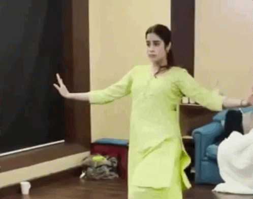 A Woman In A Yellow Dress Is Dancing In A Room With A Blue Chair . GIF