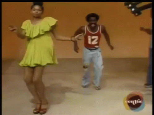 A Woman In A Yellow Dress Is Dancing Next To A Man In A Red Shirt With The Number 12 On It . GIF