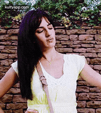 A Woman In A Yellow Dress Is Standing In Front Of A Brick Wall With Her Hands On Her Hips . GIF