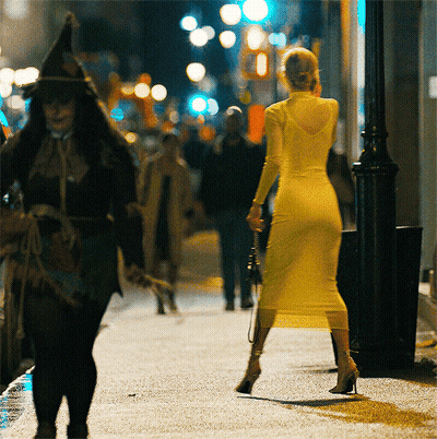A Woman In A Yellow Dress Is Walking Down A Street At Night GIF