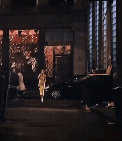 A Woman In A Yellow Dress Is Walking Down A Street In Front Of A Building With A Sign That Says Home Depot GIF
