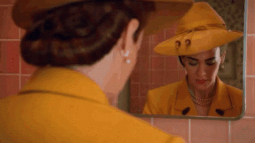 A Woman In A Yellow Hat Is Looking At Herself In A Mirror . GIF