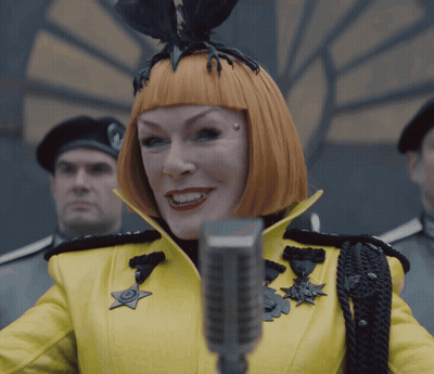 A Woman In A Yellow Jacket Is Holding A Microphone In Front Of A Crowd GIF