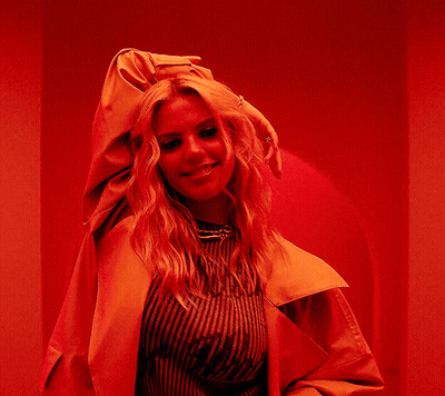 A Woman In A Yellow Jacket Is Standing In Front Of A Red Wall And Smiling . GIF