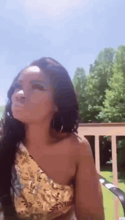 A Woman In A Yellow One Shoulder Top Is Sitting On A Chair On A Deck . GIF