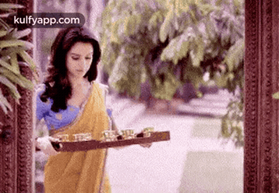 A Woman In A Yellow Sari Is Holding A Wooden Tray With Glasses On It . GIF