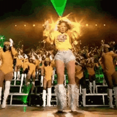 A Woman In A Yellow Shirt And Shorts Is Dancing On A Stage In Front Of A Crowd Of People . GIF