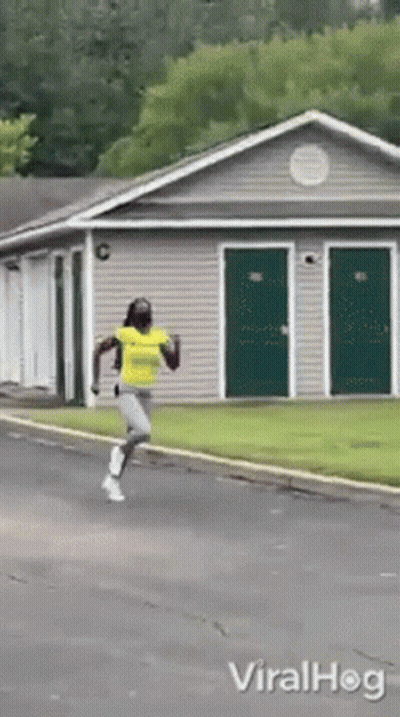 A Woman In A Yellow Shirt Is Running In Front Of A Building With Green Doors GIF